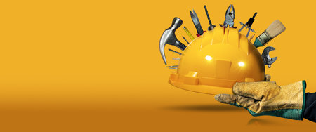 Manual worker with protective work gloves holding a yellow safety helmet with many work tools, on a yellow and orange background with copy space and reflections.の写真素材