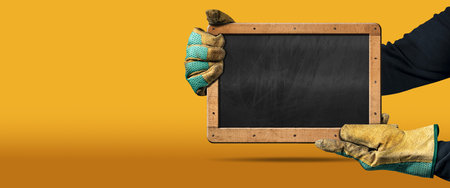Manual worker with protective work gloves holding a blank blackboard with wooden frame, on a yellow and orange background with copy space.の写真素材