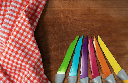 Close-up of set of six colored table knives (green, blue, purple, red, orange, yellow,) on a wooden cutting board with red and white checkered tablecloth and copy space.の写真素材