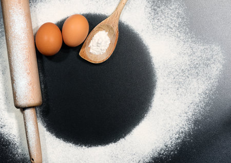 Baking ingredients on a black table with copy space, circle shape. Wooden rolling pin and spoon with white flour and two eggs.の写真素材
