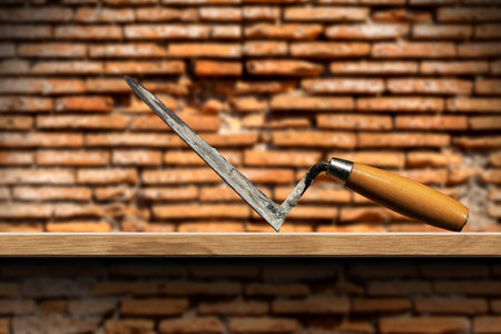 Close-up of a dirty concrete trowel with wooden handle above a wooden plank with a blurred brick wall in the background.の写真素材