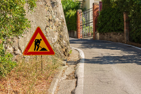 Triangular orange, red and black road sign of men at work or work in progress, by the side of a road under maintenance (bad condition). Italy, Europe.の写真素材