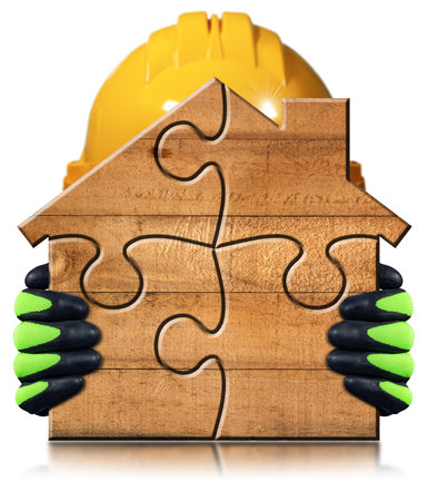 Manual worker with protective work gloves and orange hardhat holding a small wooden house formed from jigsaw puzzle pieces. Isolated on white background. Construction industry concept.の写真素材