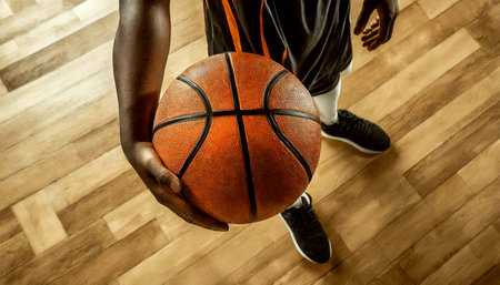 Close-up of a hand of a basketball player holding an orange and black basketball ball on the wooden parquet of a basketball court. Generative Ai.の素材