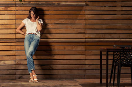 Beautiful hipster woman leaning on wooden fence wearing white t-shirt and jeansの写真素材