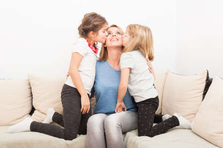Happy blonde mother with young daughters kissing as family love concept at home on a sofa or couch