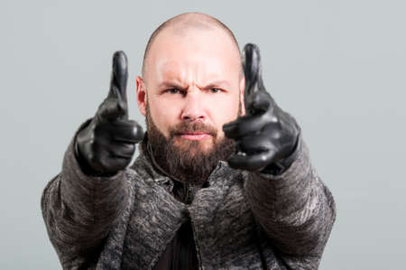 Close-up of handsome guy with beard pointing fingers gun gesture at camera on a gray background