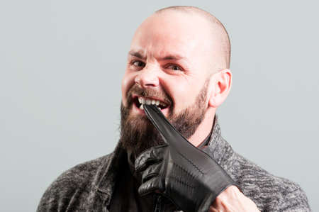 Angry bearded male biting his black glove as aggressivity concept on gray studio background