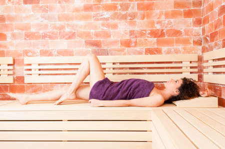 Curly young female stretched out on wooden bench relaxing in sauna roomの写真素材