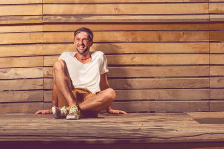 Cheerful smiling man sitting down on wooden floor with wooden fence in backgroundの写真素材