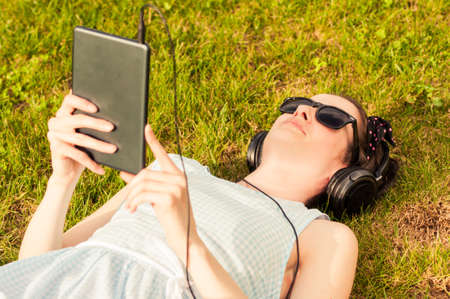 Beautiful woman holding digital tablet in park on sunny day and lying on grass as outdoor relaxation...