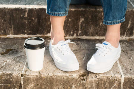 Close-up of young woman wearing blue jeans and sneakers standing near a cup of coffee outside on...