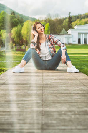 Young female in casual outfit sitting outside on wooden alley and listening music in a sunny summer dayの写真素材