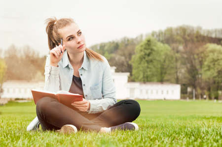 Pensive serious woman sitting outside on grass and writing in her diary with copy spaceの写真素材