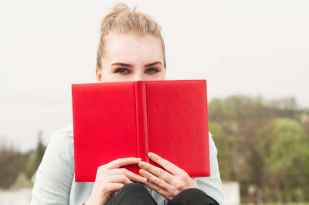 Close-up portrait of beautiful woman hiding behind red book while sitting in the parkの写真素材