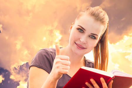Close-up portrait of attractive smiling girl reading a book and showing thumbup ok like gesture in...