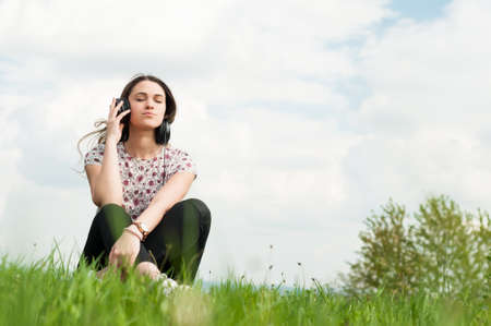 Enjoying music concept with young female sitting outside with earphones and relaxing on meadow with...