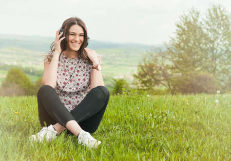 Beautiful young female with earphones outdoors enjoying music and smiling while sitting down on grass with copyspaceの写真素材