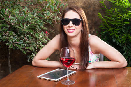 Young girl with sun glasses enjoying wine on terrace on sunny day with tablet on table and smiling with copy space areaの写真素材