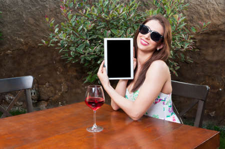 Woman wearing sun glasses showing blank display of tablet and having glass of wine outside on terrace table with copy text spaceの写真素材