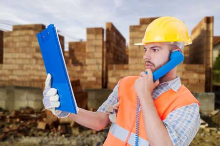 Professional constructor reading something  from clipboard notes and talking on telephone