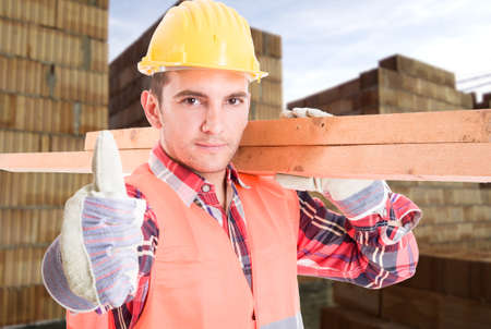 Portrait of young builder showing thumb up sign and holding pieces of wood on shoulder as renovation...