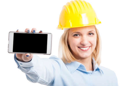 Selective focus of female engineer wearing helmet showing smartphone with blank screen and smiling isolated on white background with copy text spaceの写真素材