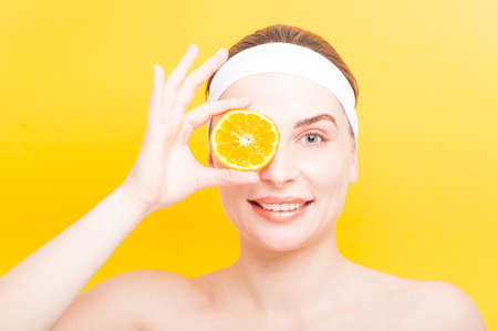 Young jouyful woman holding an piece of orange on her right eye on yellow backgroundの写真素材