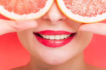 Toothy smile of beautiful woman in closeup with two slices of organic grapefruit before her eye on red backgroundの写真素材
