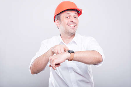 Portrait of architect wearing  hardhat showing wrist watch like being late and smiling on grey backgroundの写真素材