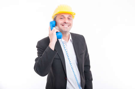 Portrait of architect talking on blue big telephone receiver wearing yellow hardhat and smiling isolated on white backgroundの写真素材
