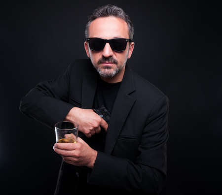 Male gangster removing his gun from jacket pocket while he is drinking scotch
