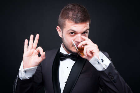 Elegant young man in suit drinking a glass of alcohol and indicate ok sign