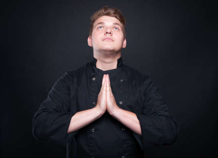 Talented young chef with palms clasped praying and looking up isolated on balck backgroundの写真素材