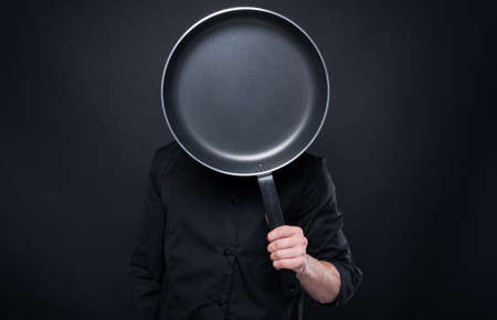 Male cook in uniform holding cooking pan in front of his face isolated on black backgroundの写真素材