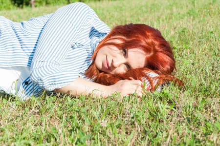 Beautiful red haired woman lying on grass outdoos relaxing on sunny dayの写真素材