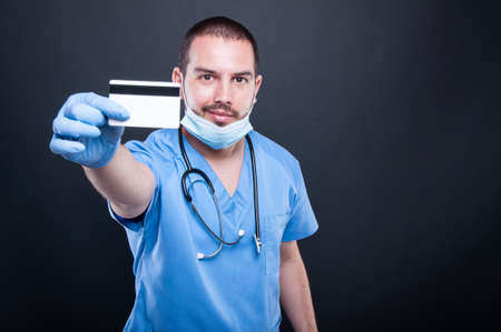 Doctor wearing scrubs and mask showing credit card smiling on black background with copy text spaceの写真素材