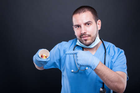 Doctor wearing scrubs pointing cup with pills and smiling on black background with copypsace advertising areaの写真素材