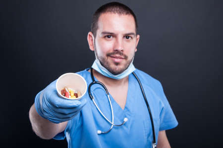 Selective focus of doctor wearing scrubs showing cup with pills and smiling on black backgroundの写真素材