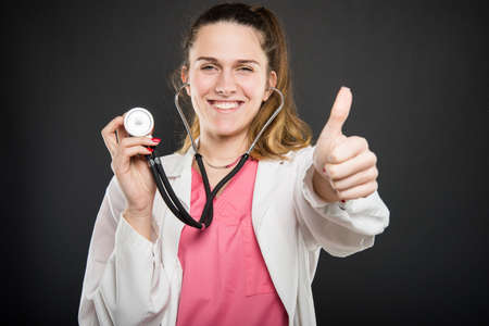 Attractive female doctor  holding stethoscope showing like gesture and smiling on black backgroundの写真素材