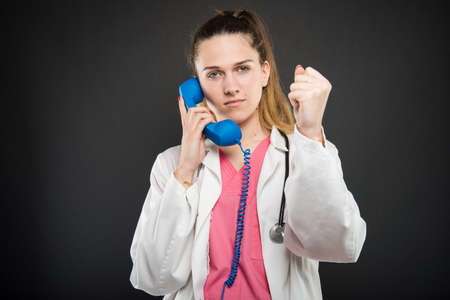 Female doctor talking at blue telephone receiver showing fist like winning on black background with copyspace advertising areaの写真素材