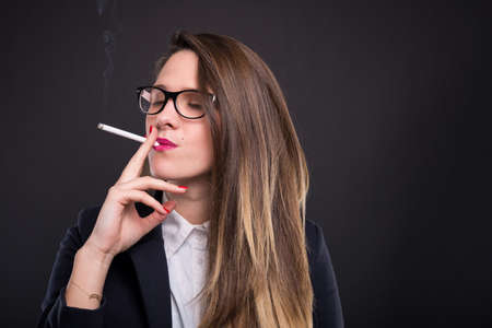 Elegant manager girl wearing glasses and smoking cigarette on dark background with copyspaceの写真素材