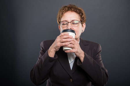 Business senior lady enjoying takeaway cup of coffee with eyes closed on black background with...