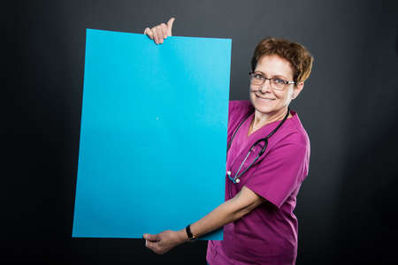 Portrait of senior lady doctor showing big blue board and smiling on black background with copyspace advertising areaの写真素材