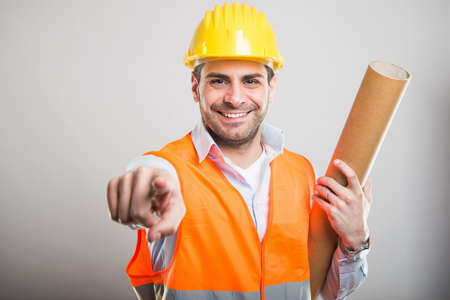 Portrait of young architect holding blueprints pointing camera and smiling on gray backgroundの写真素材