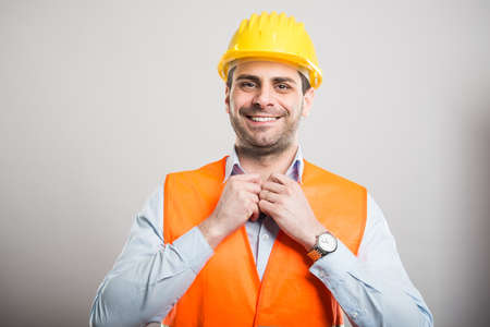 Portrait of young architect arranging his shirt wearing hardhat and smiling on gray backgroundの写真素材