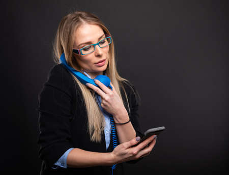 Business lady wearing blue glasses talking at blue receiver holding smartphone on black background with copyspace advertising areaの写真素材