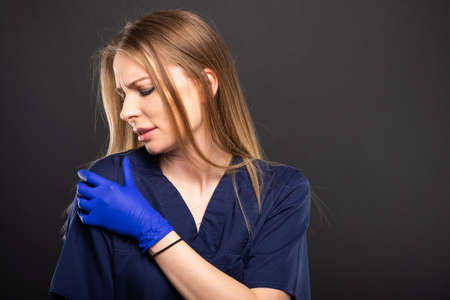 Beautiful female doctor wearing scrubs making shoulder pain gesture on black background with...
