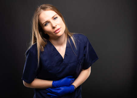 Beautiful female doctor wearing scrubs making stomach ache gesture on black background with...