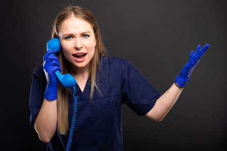 Female doctor wearing scrubs yelling at telephone receiver and gesturing on black background